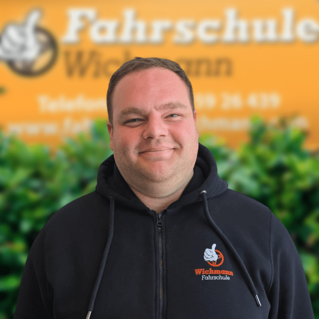 Kilian-Fahrlehrer-Fahrschule-Wichmann- - Fahrschule Wichmann Lemwerder - Autoführerschein LKW Führerschein Motorradführerschein Gunnar, Fahrlehrer der Fahrschule Wichmann Delmenhorst, lächelt vor dem gelben Firmenschild mit Logo und T-Shirt.