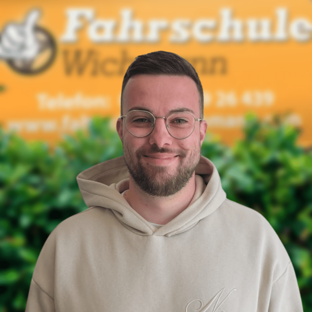 Malte-Fahrlehrer-Fahrschule-Wichmann- - Fahrschule Wichmann Lemwerder - Autoführerschein LKW Führerschein Motorradführerschein Fahrlehrer der Fahrschule Wichmann in schwarzem Logo-Pullover vor gelbem Hintergrund mit grünem Buschwerk.