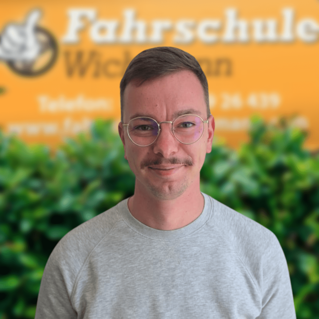 Till-Fahrlehrer-Fahrschule-Wichmann- - Fahrschule Wichmann Lemwerder - Autoführerschein LKW Führerschein Motorradführerschein Fahrlehrer Sofiane der Fahrschule Wichmann Delmenhorst vor grünem Busch und gelbem Firmenschild.