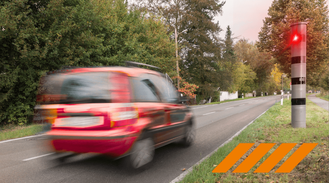 Blurred red car speeding down a two-lane road, with a red signal on a striped post and orange caution stripes in the grass at the roadside.